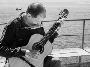 Black and white photo of Martí Batalla playing the guitar and the harmonica with the sea in the background.