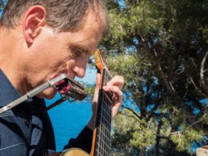 Martí Batalla playing the guitar and harmonica with pines behind that reveal the sea through their branches.