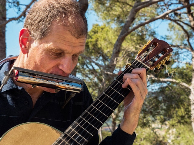 Close-up of Martí Batalla playing the guitar and harmonica with pine trees in the background.