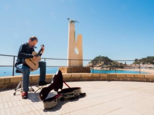 Martí Batalla in profile playing the guitar and harmonica next to the Albatross Monument in the Tossa de Mar viewpoint.