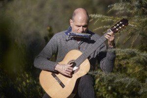 Imagen de Martí Batalla tocando la guitarra y la harmónica sentado entre árboles