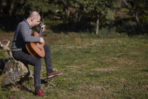 Imagen de Martí Batalla sentado en una piedra en un prado tocando la guitarra y la harmónica con un bosque al fondo